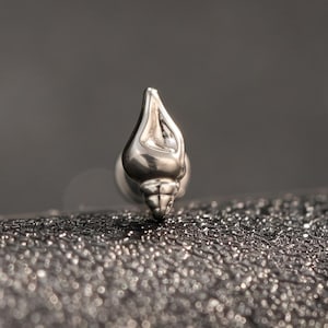 May include: A small, silver-toned metal stud earring shaped like a spiral seashell. The piece features a polished, reflective surface with a pointed top and a rounded base, designed as a singular decorative element for body piercing jewelry.
