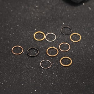 May include: A collection of ten small, circular metal rings in various finishes, including gold, silver, rose gold, and black. These thin, continuous hoops are arranged in a scattered pattern, showcasing a variety of metallic tones.