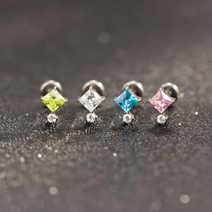 May include: Four silver stud earrings with square-cut gemstones in lime green, white, blue, and pink. Each earring features a small, round, clear gemstone below the square stone. The earrings are displayed on a dark, glittery surface.