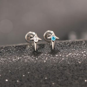 Puede incluir: Un par de pendientes de botón con forma de estrella de cuatro puntas. Un pendiente presenta una piedra preciosa redonda transparente e incolora en el centro, mientras que el otro tiene una piedra redonda de color azul brillante, ambos con base plana.