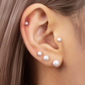 Könnte beinhalten: Nahaufnahme eines Ohrs, das mit fünf kleinen, runden, weißen Perlenohrringen geschmückt ist. Die Perlen sind unterschiedlich groß und erzeugen einen zarten und eleganten Look. Die Ohrringe sind im Ohr eingesetzt und zeigen einen klassischen Schmuckstil.