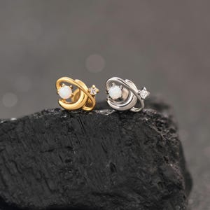 Könnte beinhalten: Zwei Ohrstecker in Planetenform mit Ringen, einer in Goldoptik und einer in Silberoptik. Jeder Ohrstecker hat in der Mitte einen runden, weißen, opalartigen Stein und einen kleinen, klaren, facettierten Stein am äußeren Ring des Planetendesigns.