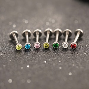 May include: A collection of seven silver-colored labret studs, each featuring a different colored gemstone. The gemstones include yellow, blue, pink, yellow, green, white, and red. The studs are arranged in a row against a dark, glittery background.