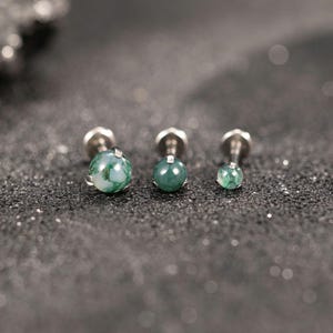 Può includere: Tre orecchini a lobo con pietre rotonde di agata muschiata verde lucida incastonate in perni di metallo color argento. Ogni orecchino presenta un motivo marmorizzato unico con inclusioni verde scuro e bianco traslucido.