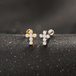 May include: Two cross-shaped stud earrings, one in gold tone and one in silver tone, each featuring five clear, round-cut stones arranged in a cross pattern. The earrings have a flat back post design, showcasing a metallic finish and clear stones.