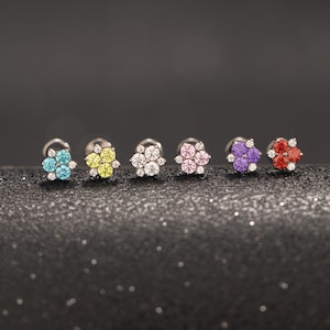 Puede incluir: Seis pendientes de botón con forma de flor. Cada pendiente presenta un grupo de gemas de colores en tonos azul, amarillo, blanco, rosa, morado y rojo, rodeadas de piedras blancas más pequeñas. Los pendientes están engarzados en postes de plata.