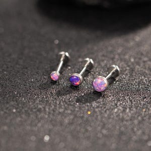 May include: Three silver labret studs with iridescent, oval opal stones. The studs vary in size, with the largest on the right. The opals display shades of purple, pink, and orange. The background is a dark, textured surface.