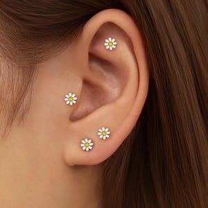 Puede incluir: Cuatro pendientes de botón de margarita con pétalos blancos y centros amarillos se muestran en una oreja. Los pendientes son pequeños y delicados, con un diseño floral sencillo. Los pendientes son un accesorio ligero y brillante.