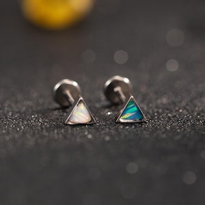 Könnte beinhalten: Ein Paar silberne Dreiecks-Ohrstecker. Ein Ohrring hat ein weißes und schillerndes Design, der andere ein blaues und grünes schillerndes Design. Die Ohrringe sind vor einem dunklen, glitzernden Hintergrund platziert.