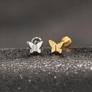 Peut inclure: Deux boucles d'oreilles à clous en forme de papillon, une argentée et une dorée. Elles sont posées sur une surface sombre et texturée avec un arrière-plan flou. Les ailes des papillons sont détaillées.