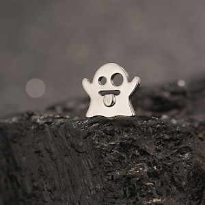 Puede incluir: Un dije plateado con forma de fantasma con un diseño juguetón. El dije presenta un ojo redondo, un ojo redondo más pequeño y una lengua afuera. El dije está sobre un fondo oscuro y texturizado.