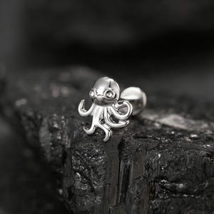 May include: A silver-colored octopus-shaped earring. The earring features a detailed design of an octopus with curved tentacles and a smiling face. The earring has a post and a round backing. The background is a dark, textured surface.