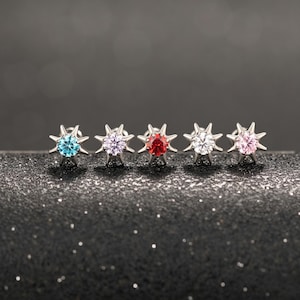 May include: Five starburst-shaped stud earrings with colorful gemstones. The earrings feature a silver setting and are adorned with a variety of gemstones, including blue, purple, red, white, and pink. The earrings are displayed on a dark, glittery surface.
