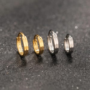 Puede incluir: Cuatro pendientes de aro en tonos dorados y plateados. Los pendientes tienen un diseño facetado, con dos aros dorados y dos plateados. Se exhiben sobre una superficie oscura y moteada, mostrando su brillo metálico y textura geométrica.