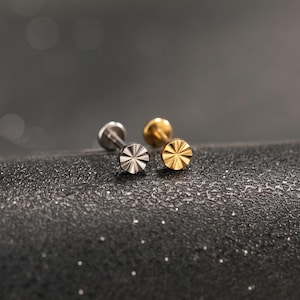 May include: Two flat-back stud earrings, one in a silver tone and one in a gold tone. Each stud features a circular head with a sunburst or starburst pattern of etched lines radiating from the centre, creating a textured, reflective surface.