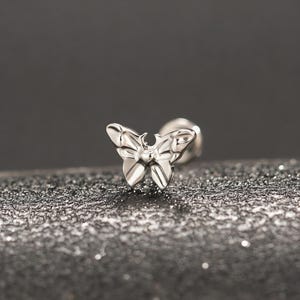 Könnte beinhalten: Ein kleiner, silberner Ohrstecker in Schmetterlingsform. Der Schmetterling hat detaillierte Flügel- und Körpermerkmale. Der Ohrringstift ist hinter dem Schmetterling sichtbar. Der Hintergrund ist eine dunkle, strukturierte Oberfläche.