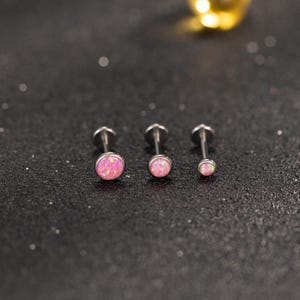 Può includere: Tre piercing in argento per labbra o cartilagine con pietre rosa opalescenti. I piercing sono disposti in fila, con il più grande a sinistra e di dimensioni decrescenti. Lo sfondo è una superficie scura e strutturata con piccoli riflessi scintillanti.