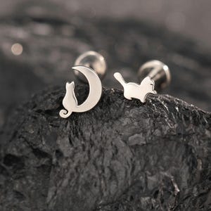 May include: A pair of silver-toned stud earrings featuring a cat silhouette sitting inside a crescent moon shape and a separate cat silhouette in a walking pose. The earrings have a polished metallic finish and are displayed on a dark rock.