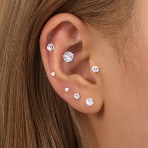 May include: An ear with multiple piercings, each featuring a clear, round gemstone. The gemstones are of varying sizes, creating a layered effect. The ear is set against a backdrop of light brown hair.