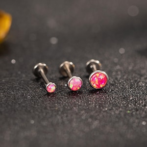 Può includere: Tre piercing per labbra o cartilagine color argento con pietre rosa simili all'opale. I borchie sono disposti in fila, con le pietre di opale di dimensioni variabili. Lo sfondo è una superficie scura e glitterata.
