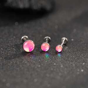 Könnte beinhalten: Drei silberfarbene Lippen- oder Ohrstecker mit schillernden rosa und orangefarbenen runden Oberteilen. Die Stecker sind in einer Reihe angeordnet und verkleinern sich von links nach rechts. Der Hintergrund ist eine dunkle, strukturierte Oberfläche.
