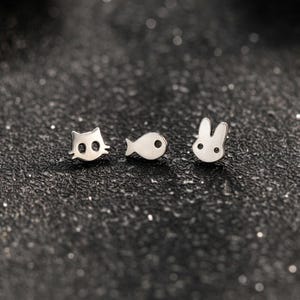 May include: Three silver-toned metal stud earrings featuring distinct shapes: a cat head with cut-out eyes, a small fish with a circular eye, and a rabbit head with long ears and two small circular cut-outs for eyes, all with a polished finish.