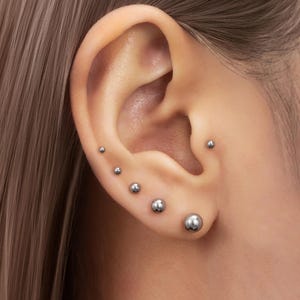 Può includere: Primo piano di un orecchio con diversi orecchini a sfera d'argento di varie dimensioni. Gli orecchini sono distanziati in modo uniforme lungo la cartilagine e il lobo dell'orecchio, creando un effetto a strati. Il tono della pelle è chiaro, i capelli sono castano chiaro.