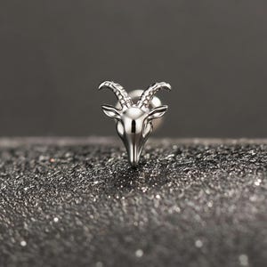 May include: A silver-colored goat head stud earring. The earring features a detailed depiction of a goat's head with curved horns and a pointed chin. The earring is set against a dark, textured background.