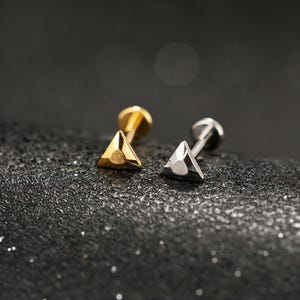 May include: Two triangle-shaped stud earrings. One is gold-colored, the other is silver-colored. Both earrings have a flat back and a triangular front. The earrings are displayed on a textured, dark surface.