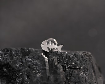 May include: A silver-colored fish bone stud earring with a small, clear gem on the head. The earring is set against a dark, textured background, highlighting its delicate design and metallic sheen.