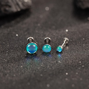 May include: Three silver-toned stud earrings with iridescent, teal-colored, round gemstones. The earrings are arranged in a row, showcasing varying sizes. The gemstones have a subtle, color-shifting effect, reflecting shades of blue and green.