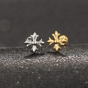 Könnte beinhalten: Zwei Ohrstecker mit Kreuz-Design. Ein Ohrring ist silberfarben, der andere goldfarben. Sie liegen auf einer strukturierten, dunkelgrauen Oberfläche.