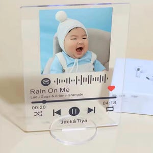 Könnte beinhalten: Transparenter Acryl-Fotorahmen mit einem Baby, das einen weißen Hut trägt, mit dem Songtitel "Rain On Me" und einer Musikwellenform-Grafik. Der Rahmen enthält die Namen "Jack & Tiya" und ein Herzsymbol.