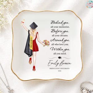 May include: A white ceramic trinket dish with a gold rim, featuring a graduation-themed illustration and text. The illustration depicts a graduate in a red dress and cap. Text includes the name "Emily Brown" and "Princeton University 2026".