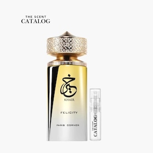 May include: A perfume bottle with a gold-colored textured cap and a clear glass body. The bottle has a black and white design with the word "KHAIR" in black script. A small sample bottle is next to it, with the text "THE SCENT CATALOG".