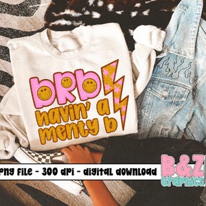 Puede incluir: Sudadera crema con el texto "brb havin' a menty b" en rosa y dorado, con caras sonrientes y gráficos de rayos. También se ve una chaqueta vaquera con detalles de llamas y un par de zapatillas a cuadros.