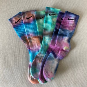 May include: Five pairs of tie-dye socks with a black Nike swoosh logo on each sock. The socks are in shades of blue, green, and purple.