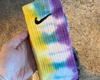May include: A pair of tie-dye socks with a black Nike swoosh logo. The socks are yellow, blue, green, and purple.
