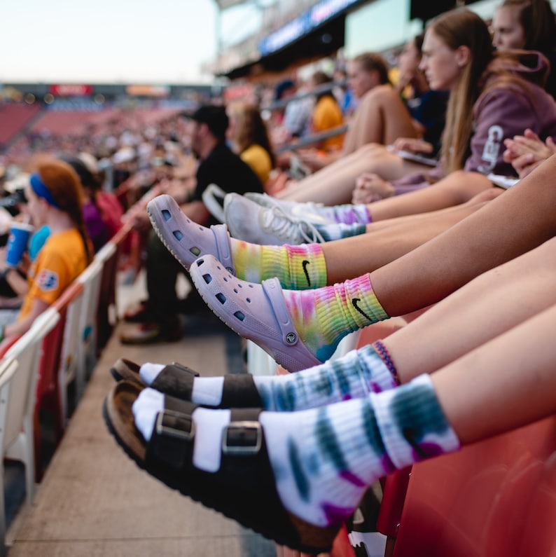May include: Close-up of four people wearing colorful tie-dye socks and various footwear, including sandals and Crocs, while sitting in stadium seats.