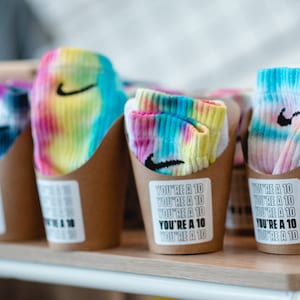 May include: Four brown paper cups with tie-dye socks inside. Each cup has a sticker that says "You're a 10" repeated multiple times.