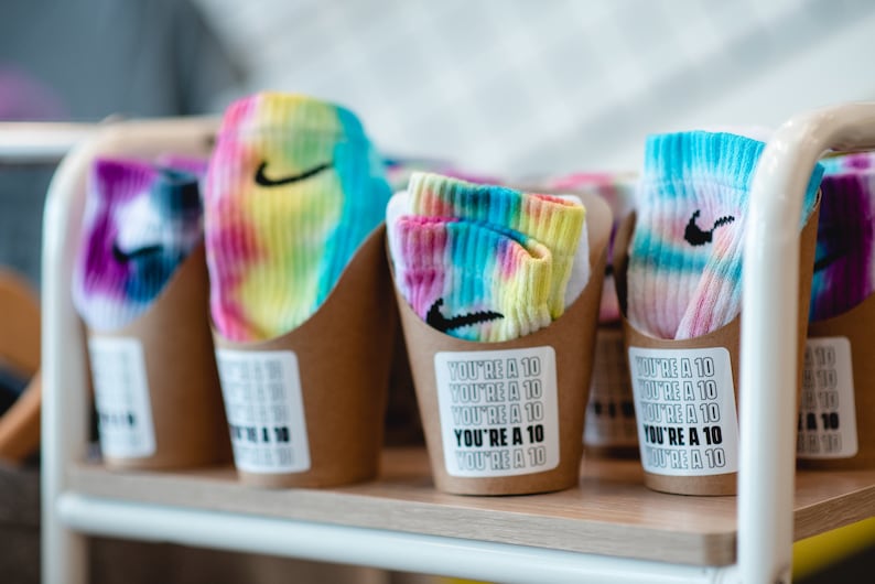 May include: Four brown paper cups with tie-dye socks inside. Each cup has a sticker that says "You're a 10" repeated multiple times.