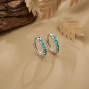Puede incluir: Un par de pendientes de aro plateados con una fila de pequeñas piedras turquesas brillantes. Los pendientes se exhiben sobre una superficie marrón claro, con un trozo de madera y hojas secas en el fondo. Un diseño de joyería sencillo y elegante.