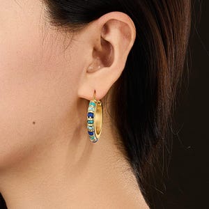 Può includere: Orecchino a cerchio dorato con un design a perline. Le perline sono in tonalità di blu, verde acqua e crema. L'orecchino è mostrato sull'orecchio di una persona, con capelli scuri sullo sfondo.