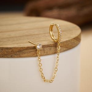 May include: A gold-toned earring featuring a small hoop encrusted with clear stones, connected by a delicate chain to a single stud earring with a clear stone. The metal has a polished finish, and the design creates a linked, dual-piercing look.