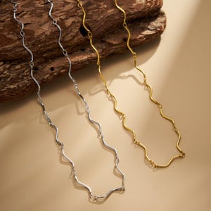 May include: Two metal chains, one silver-toned and one gold-toned, each featuring a series of interconnected wavy, serpentine links. The chains are displayed side-by-side, showcasing their distinct metallic finishes and the repeating curved pattern of the individual links that form the length of each chain.