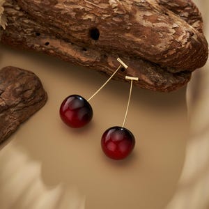Puede incluir: Un par de pendientes con forma de cereza. Cada pendiente presenta un colgante de cereza brillante, con degradado rojo y negro, suspendido de una fina barra dorada. Los pendientes están colocados sobre un fondo neutro, de color beige, con un trozo de madera.