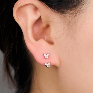 May include: Two small, silver butterfly earrings with clear, sparkling stones are displayed on an ear. The earrings are positioned one above the other, with the top earring slightly higher on the earlobe.
