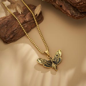 Puede incluir: Un collar dorado con un colgante detallado de polilla. El colgante tiene intrincados detalles de alas y cuerpo con acentos negros. La cadena es de estilo caja. El collar descansa sobre una superficie de tono neutro.