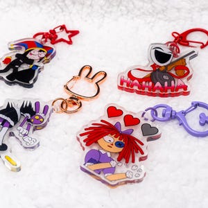 Op de afbeelding: Vijf acryl sleutelhangers met cartoon karakter ontwerpen. Eén heeft een nar, een ander een karakter met een gitaar en het woord "HAHAHA", en anderen met verschillende cartoonfiguren. Elke sleutelhanger heeft een andere kleur sluiting.