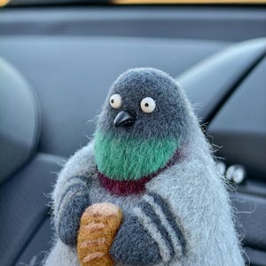 May include: A needle-felted pigeon figurine with a fuzzy grey body, a vibrant green and deep purple neck patch, and a small black beak. The pigeon has wide white eyes with black pupils and is holding a small, textured golden-brown loaf of bread.